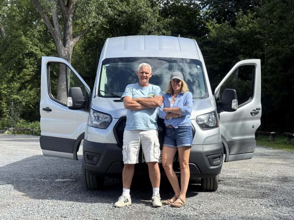 The Van Adventurers in front of the fitted van with open doors, surrounded by lush green trees, enjoying outdoor life and nature.