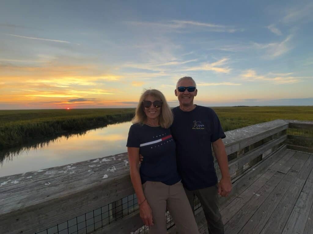 A smiling couple enjoying a sunset view at a nature reserve, standing on a wooden observation deck with a river and open sky in the background.