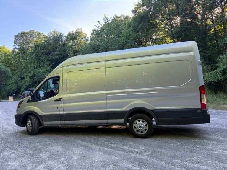 Large white cargo van parked on a gravel surface surrounded by lush green trees and foliage, under a clear blue sky.