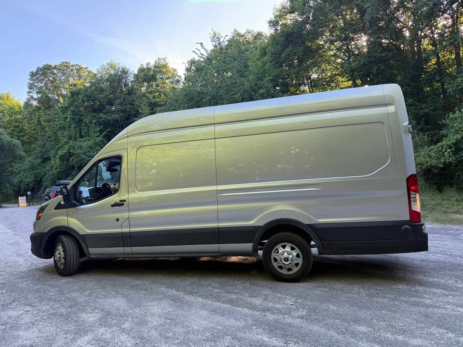 Large white cargo van parked on a gravel surface surrounded by lush green trees and foliage, under a clear blue sky.
