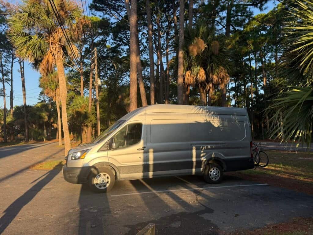 A silver van parked in a forested area with tall trees and palm trees, under a clear blue sky, during daytime.